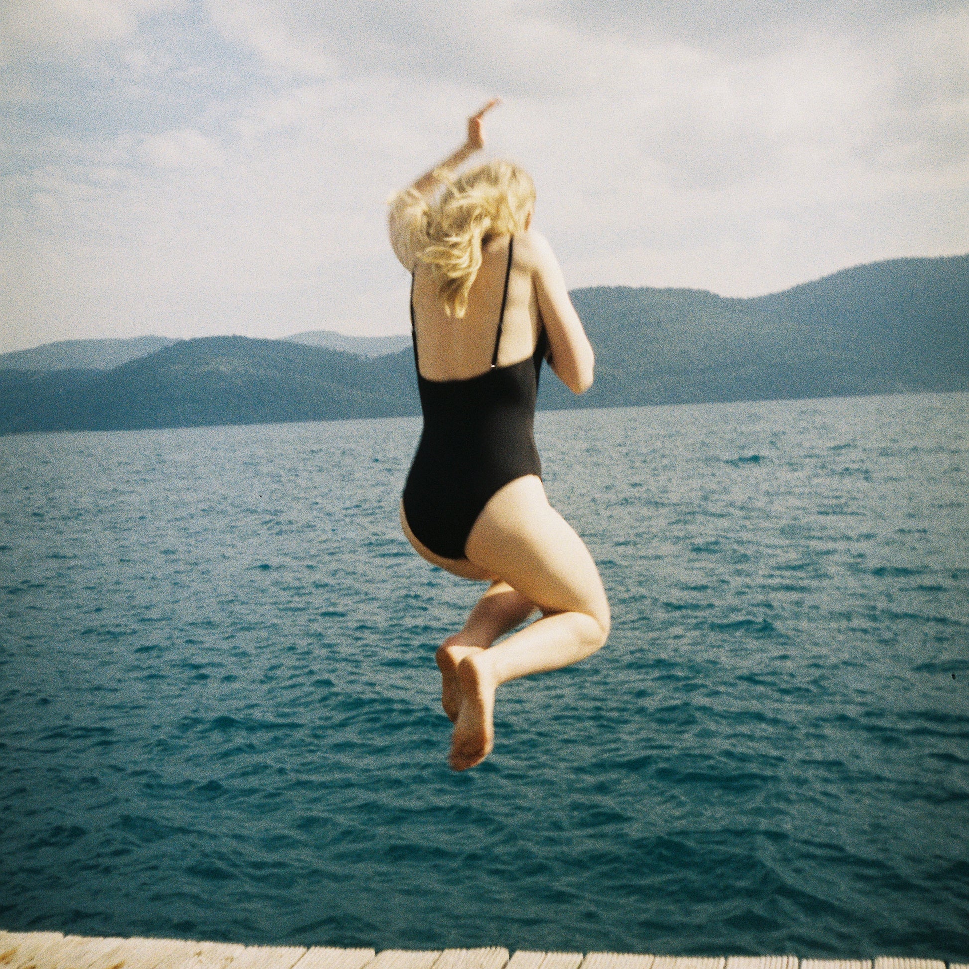 Person in a black swimsuit jumping off a wooden dock into a body of water with mountains in the background.